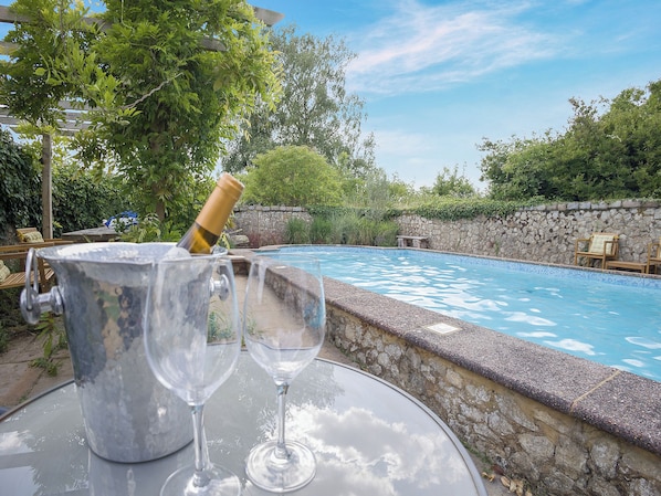 A poolside setting with a stone wall, a wooden table, a metal bucket, and two empty wine glasses.