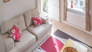 Ein Wohnzimmer mit einem beige gefärbten Sofa, zwei blumigen Kissen, einem Holztisch und Blick ins Freie durch ein Fenster.