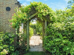 Un bâtiment en briques surmonté d’une arche en bois recouverte de verdure et de fleurs roses, qui mène à un sentier pavé de pierres.
