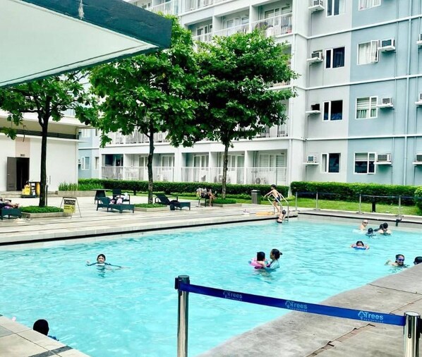 A swimming pool with people in the water, surrounded by a building with balconies and trees.
