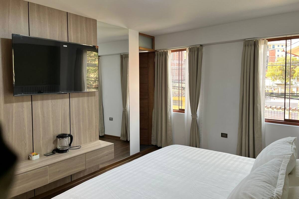 A modern bedroom with a flat-screen TV mounted on a wooden panel wall, a bed with white bedding, a bedside table with a coffee maker, and a window with curtains.
