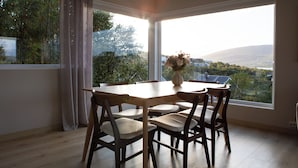 Un coin repas avec une table et des chaises en bois, un vase rempli de fleurs et une grande fenêtre donnant sur un paysage de arbres et de collines.