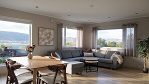 Un vaste espace de vie doté d’une grande fenêtre offrant une vue panoramique, d’un canapé d’angle gris, d’une table à manger en bois avec des chaises et d’une plante en pot.