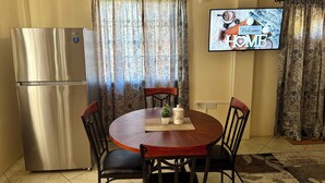 Una cocina compacta con un refrigerador de acero inoxidable, una mesa de madera redonda con cuatro sillas y un televisor de pantalla plana montado en la pared mostrando un mensaje de bienvenida.