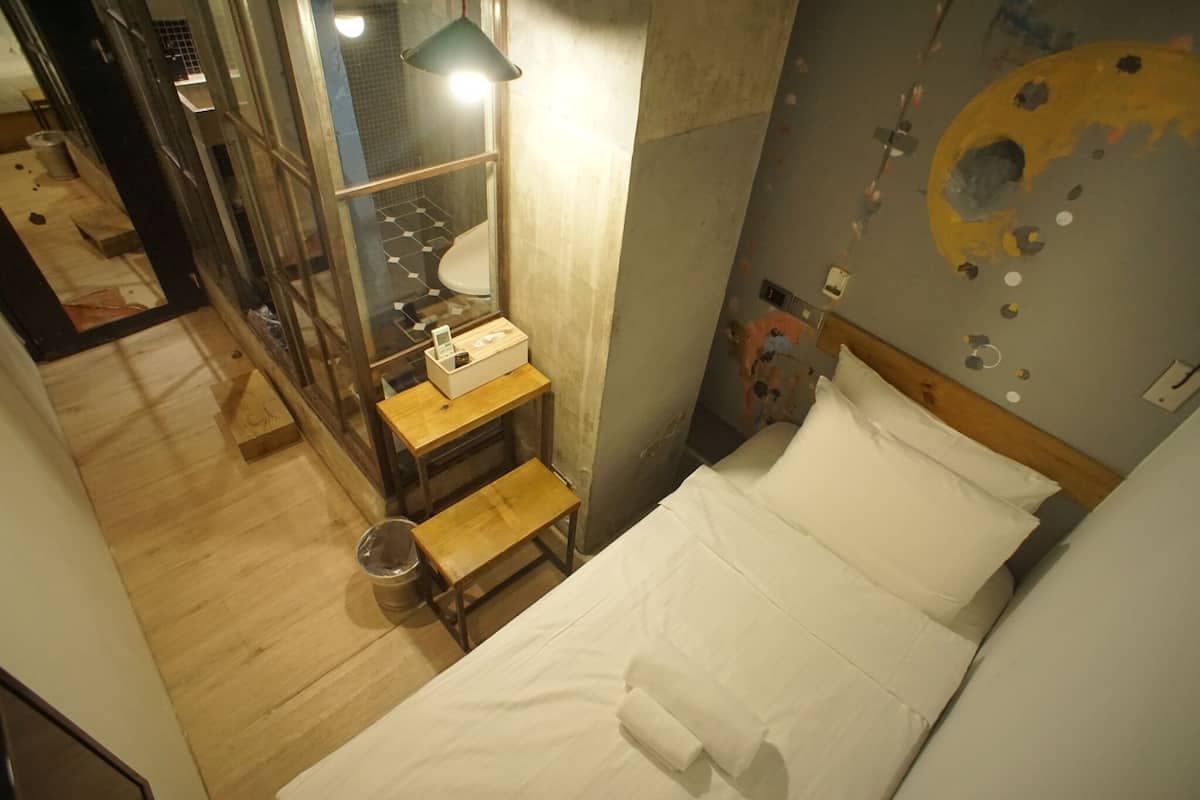 A room with a bed, a wooden bench, a small table with toiletries, and a wall with a circular design.