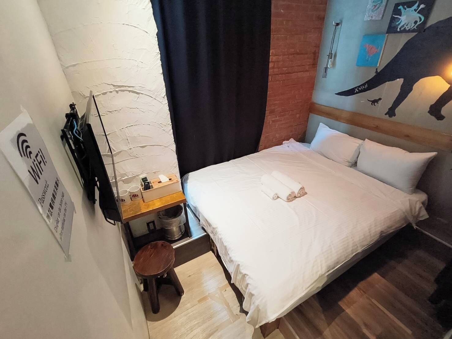 Ett hotellrum med en säng, ett litet träbord med en papperskorg, en väggmonterad skylt som visar att det finns Wi-Fi och en väggmålning.