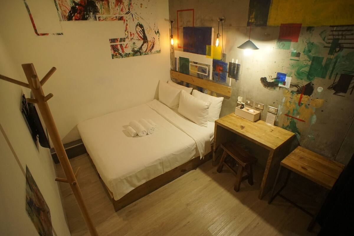 A small, cozy bedroom with a single bed, a wooden desk, and a chair. There is a wooden coat rack and a small stool. The walls are decorated with various artworks and posters.