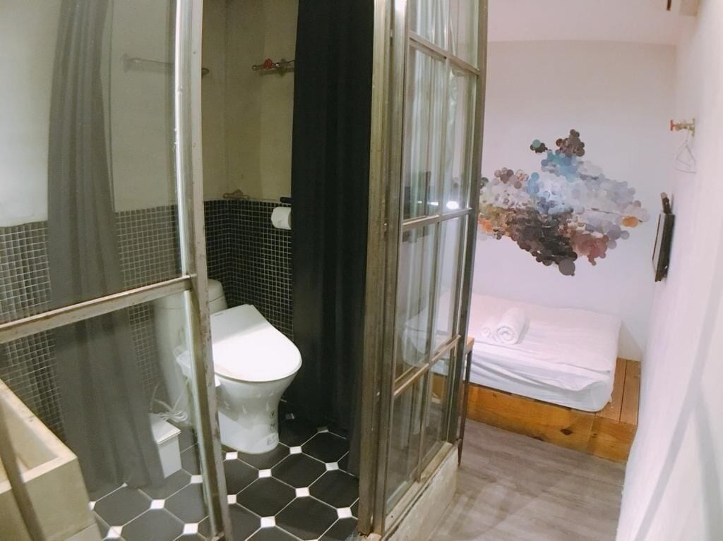 A bathroom with a glass shower enclosure, a toilet, and a bed with a white mattress and pillows.