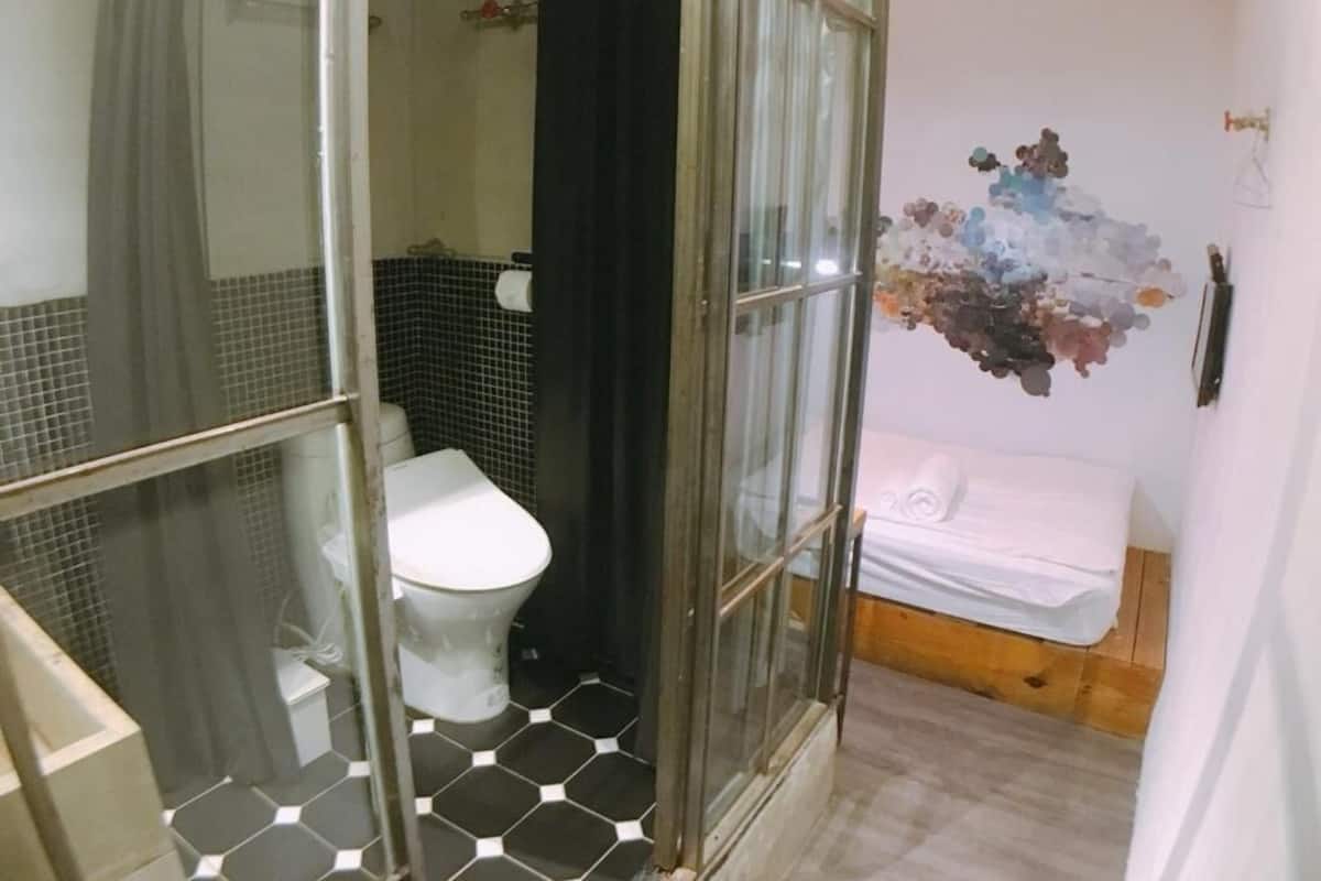 A bathroom with a glass shower enclosure, a toilet, and a bed with a white mattress and pillows.
