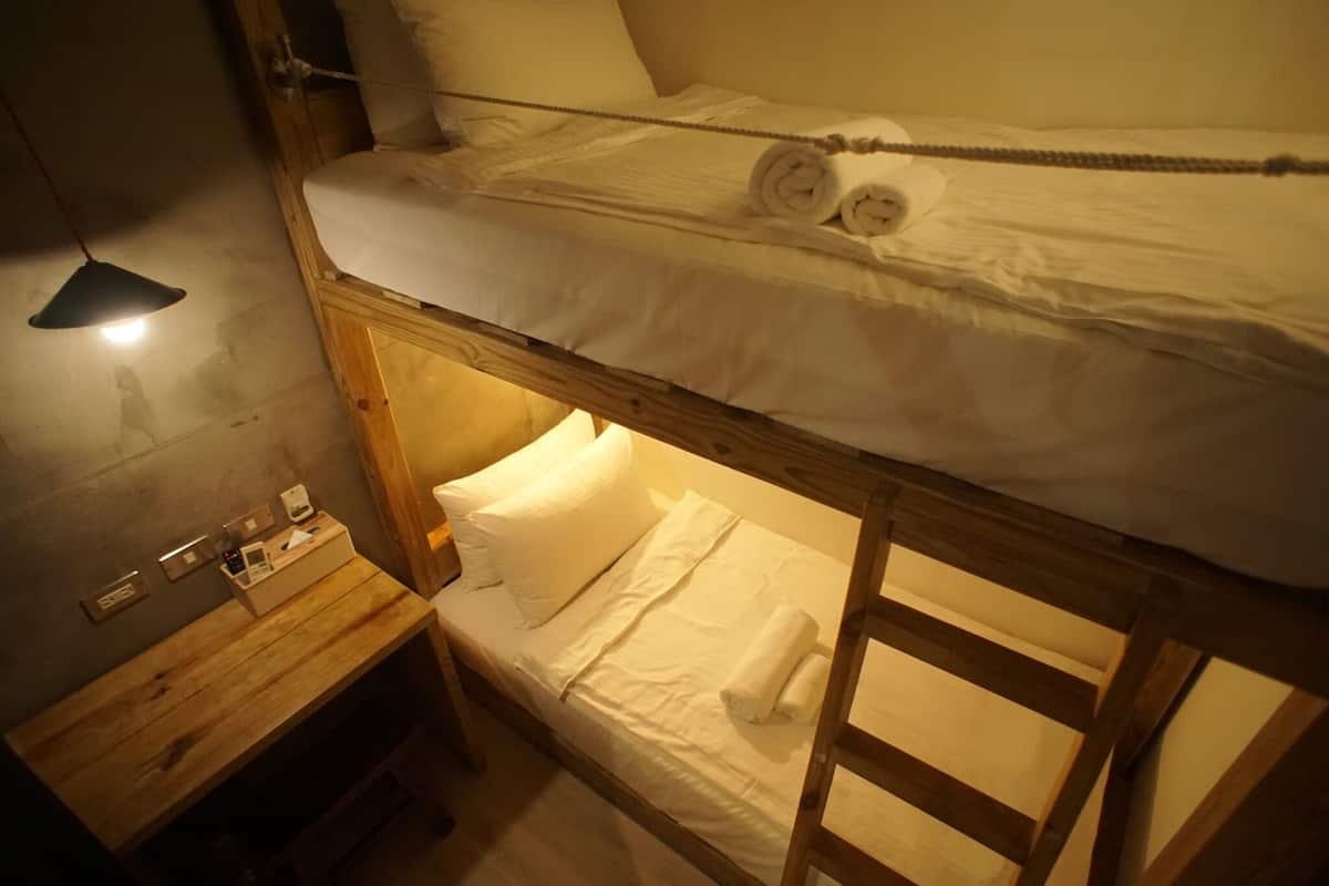 A bunk bed with a wooden frame, white bedding, and a small wooden table with a lamp and a box of toiletries.