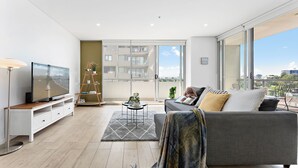 Ein modernes Wohnzimmer mit einem grauen Sofa, einem Flachbildfernseher auf einem weißen Schrank, Holzboden und einer großen Glasschiebetür, die auf einen Balkon führt.