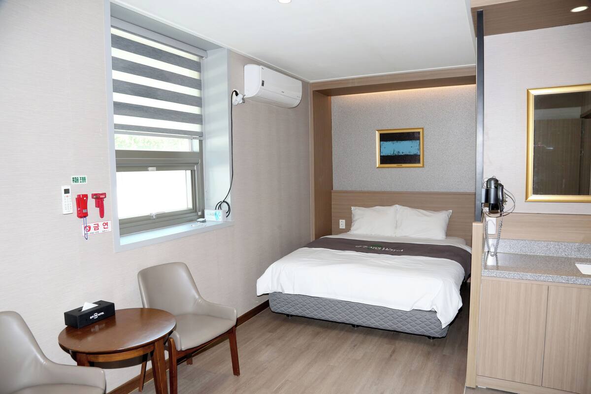 Een hotelkamer met een bed, een bureau, een stoel, een raam met jaloezieën, een airconditioner, een schilderij aan de muur en een spiegel.