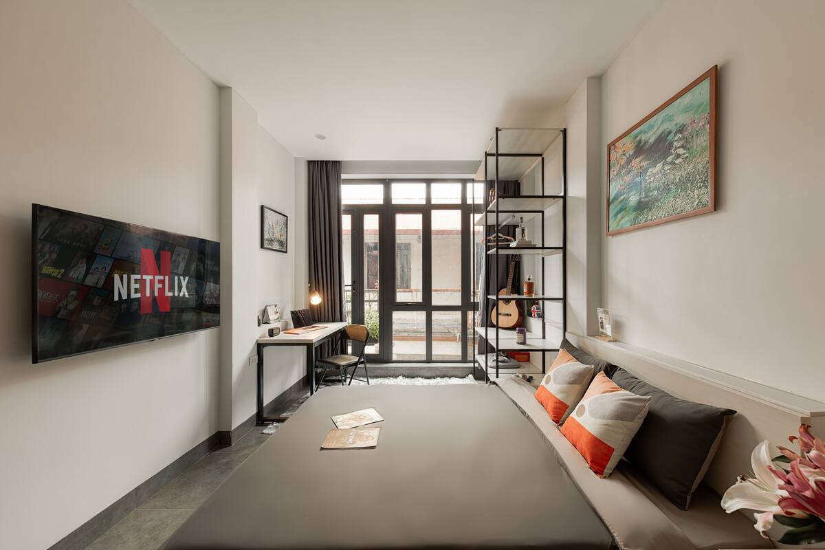 A modern hotel room with a large bed, a desk with a chair, a bookshelf, and a large window.