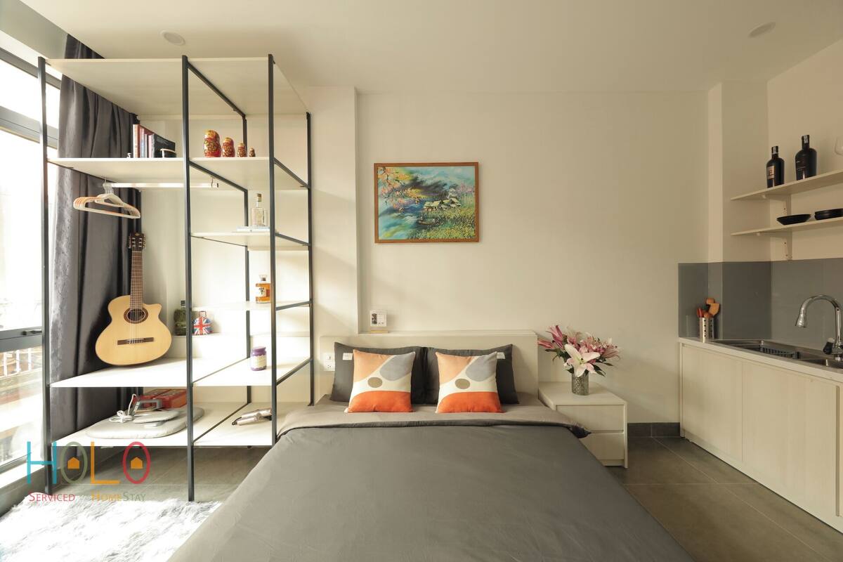 A modern bedroom with a bed, a guitar, a bookshelf, a painting, and a kitchen area.
