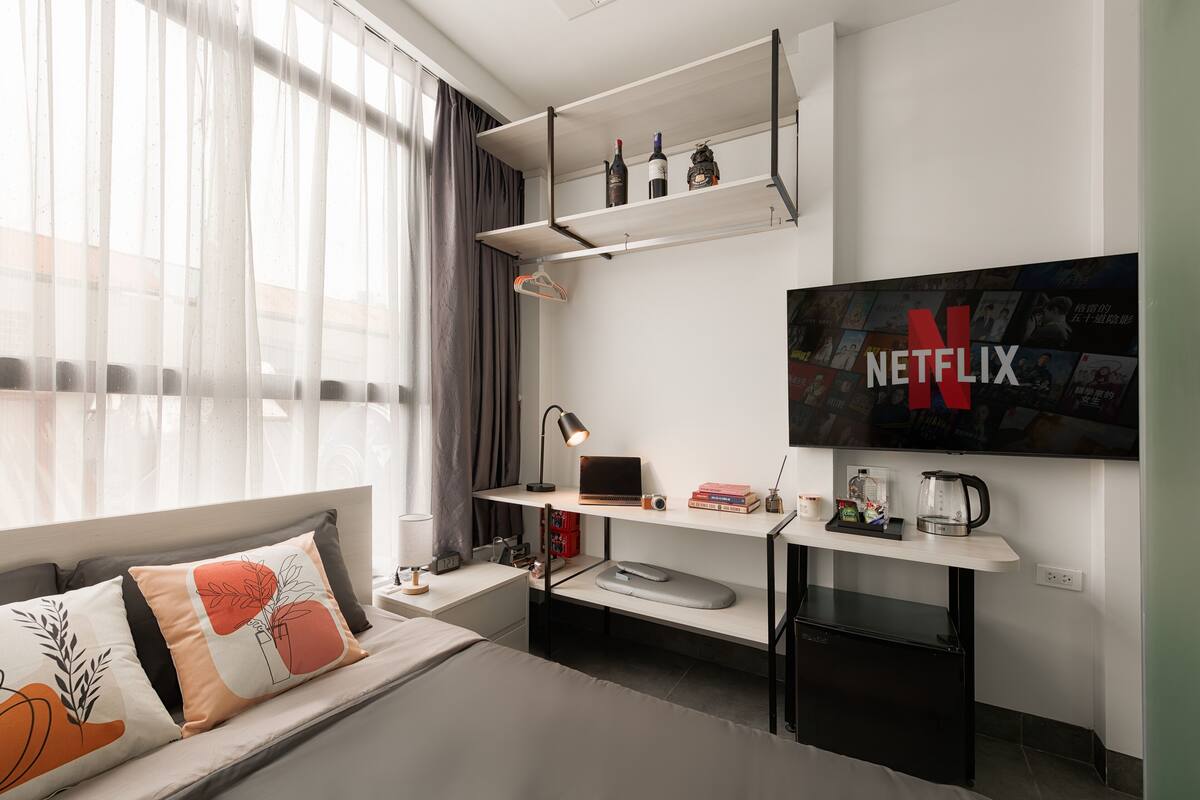 A modern hotel room with a bed, a desk with a laptop, a coffee maker, and a TV mounted on the wall displaying the Netflix logo.