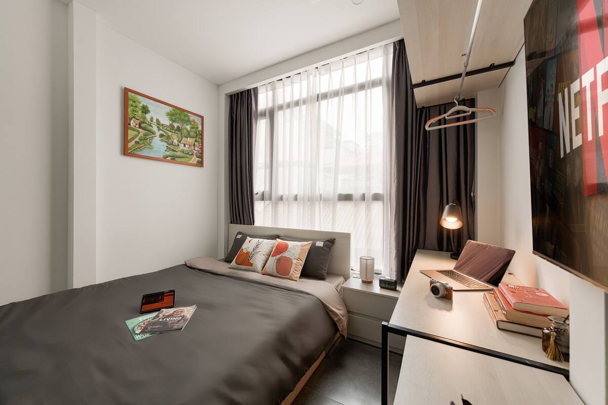 A modern hotel room with a bed, a desk with a laptop, a camera, and a magazine on the bed.