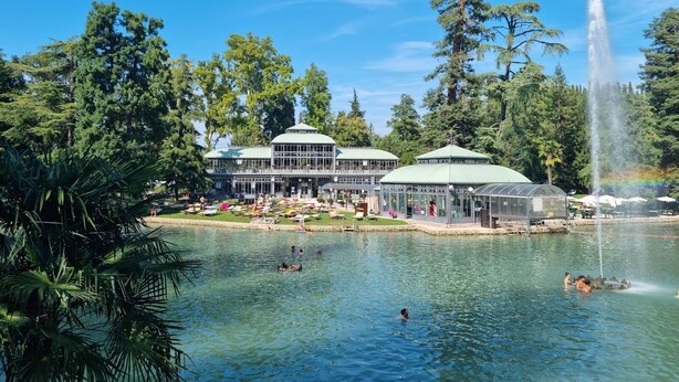 Un hotel junto al lago con varias construcciones de techo verde, una fuente y gente nadando y descansando junto al agua.