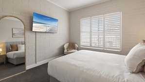 A bedroom with a large bed, a TV mounted on the wall, a chair, and a window with blinds.