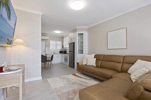 Un salon moderne avec un canapé marron, un coin repas avec une table et des chaises, une cuisine avec des meubles blancs et un luminaire au plafond.