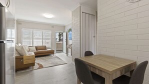 A modern living room with a sofa, a dining table with chairs, a kitchen area with a refrigerator, and a white brick wall.