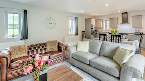 A modern living room with a brown leather sofa, grey sofa, and a wooden coffee table. There is a dining area with a table and chairs, and a kitchen with cabinets and a stove.