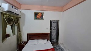 A room with a bed, a wall-mounted air conditioner, a mirror, a door with a decorative pattern, and a small picture on the wall. - Shri Yamuna Dham (Mathura)