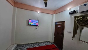 A room with a flat-screen TV mounted on the wall, a ceiling fan, an air conditioning unit, a red carpet, and a door with a sign. - Shri Yamuna Dham (Mathura)