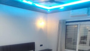 A bedroom with a bed, a nightstand, a wall-mounted light, and a window with curtains. - Madhurai Resort (Chikhaldara)