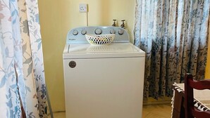 A compact, white washing machine with a front-loading door, a basket on top, and a wall-mounted power outlet.