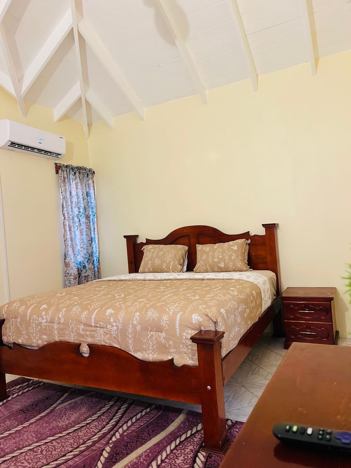 A bedroom with a wooden bed, a nightstand, a remote control, and a patterned rug.