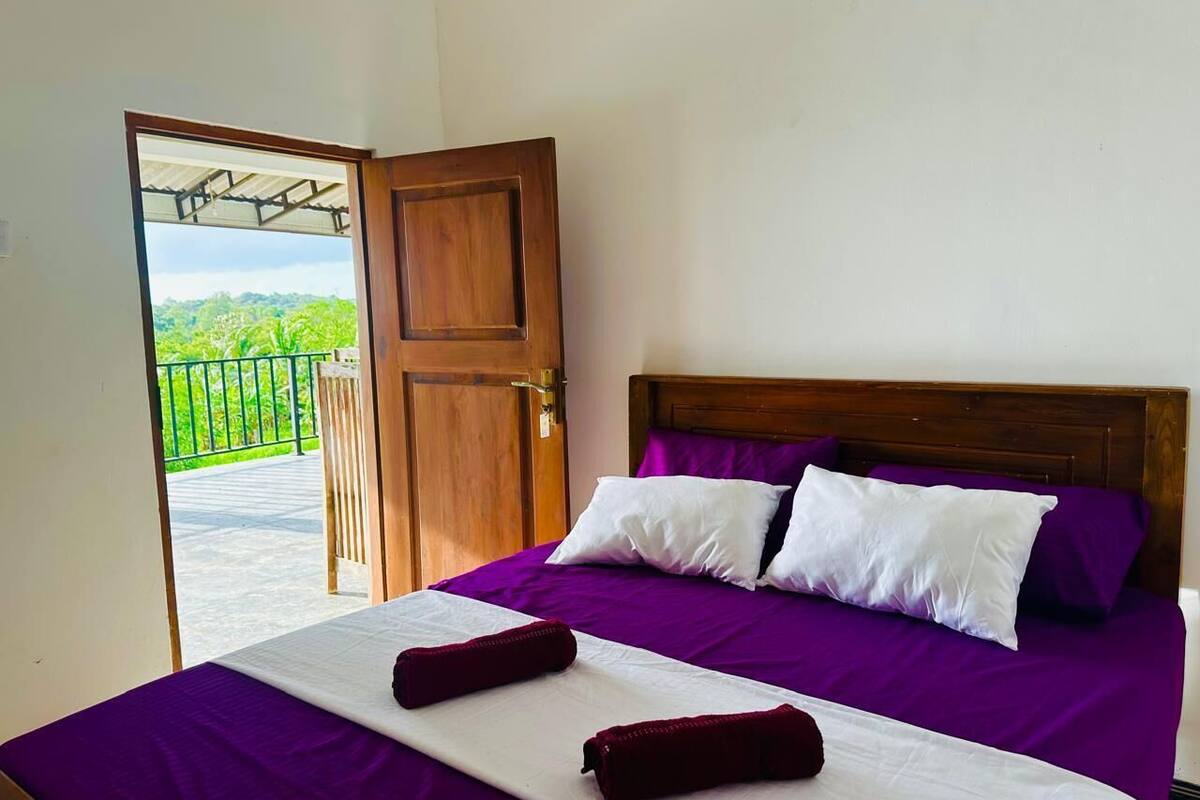 Un dormitorio con cama, ropa de cama púrpura y vistas a la vegetación por una puerta abierta.