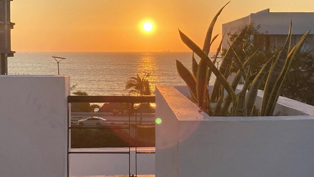 A balcony with a view of the sunset over the ocean, a white building, and some tropical plants. - Roma Suite A Malecon of Mazatlan (Mazatlan)
