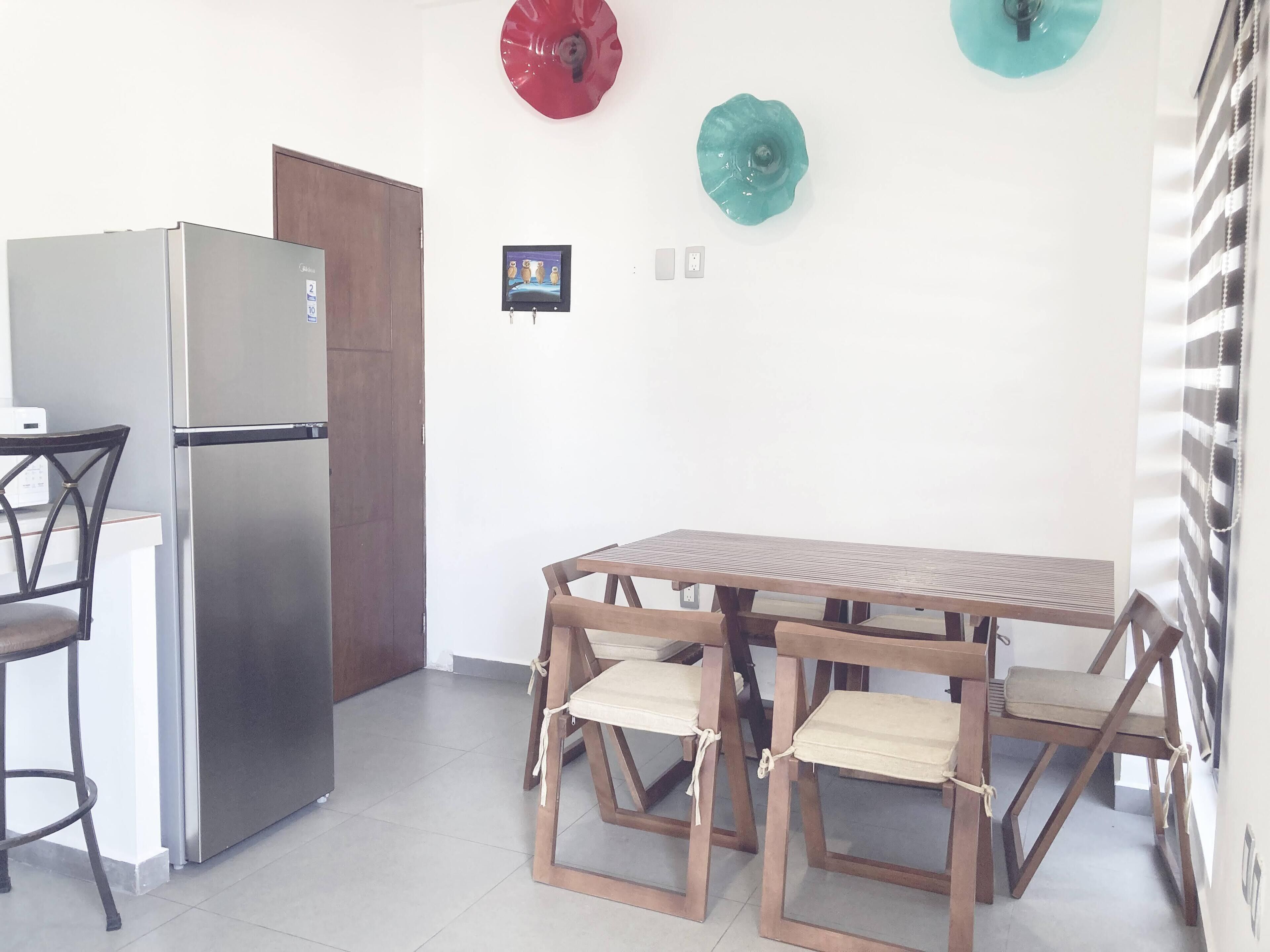 A compact kitchen with a stainless steel refrigerator, a wooden dining table with four chairs, and a door leading to another room.