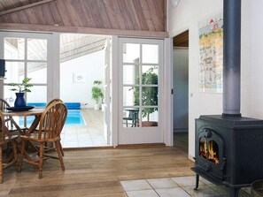 A room with a wooden floor, a dining table with chairs, a blue vase, a potted plant, a fireplace, and a view of a pool and greenery outside. - 4 Star Holiday Home in Vig (Vig)