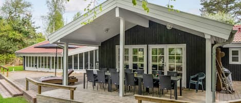 A covered outdoor seating area with tables and chairs, a wooden deck, and a view of a building with a red-tiled roof.