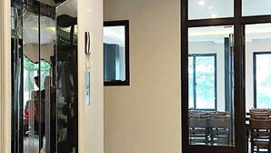 Un pasillo moderno de hotel con un ascensor de cristal, una puerta de madera y vistas a un comedor con mesas y sillas.