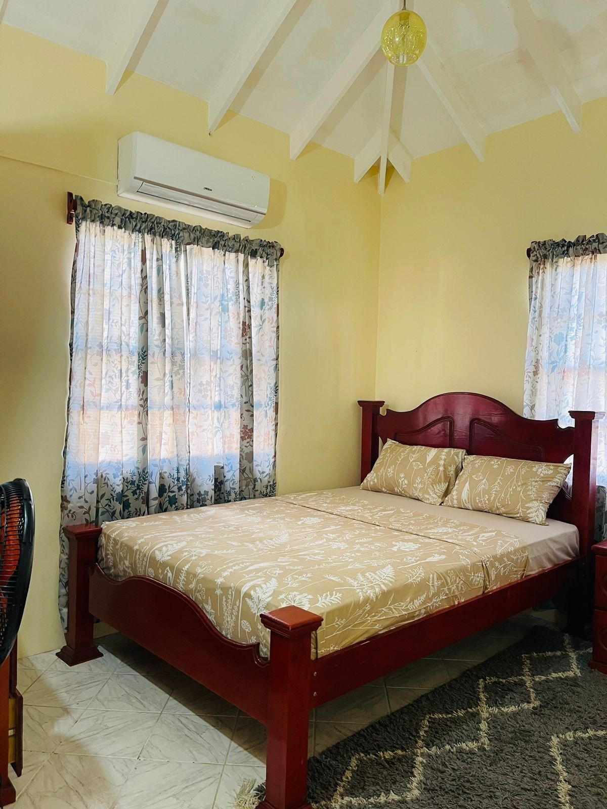 A bedroom with a wooden bed, floral curtains, an air conditioning unit, and a ceiling fan.