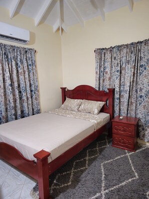 A bedroom with a wooden bed, floral curtains, a bedside table, and a rug. - Spacious 3-bedroom apartment with WiFi, AC at amazing Castle Berry (kingstown)