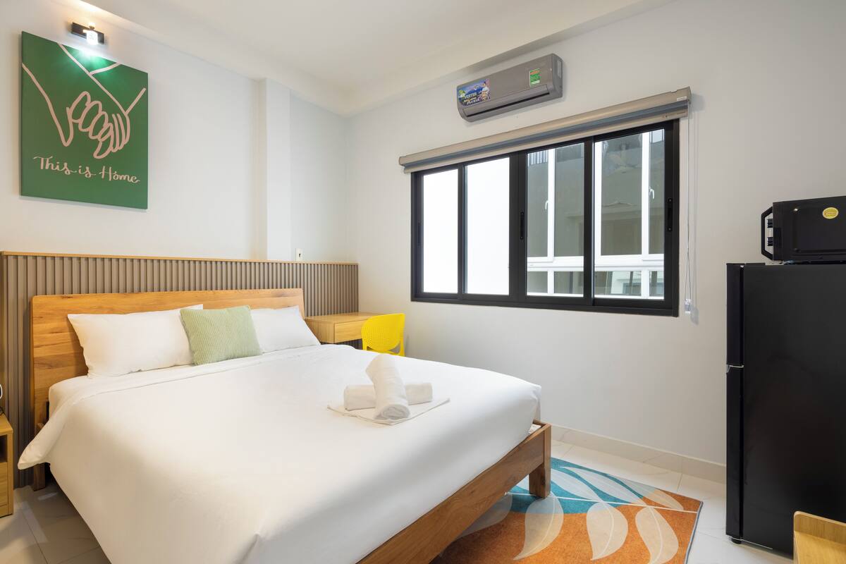 Une chambre d’hôtel avec un lit en bois, un petit bureau, une chaise, une fenêtre et un réfrigérateur.