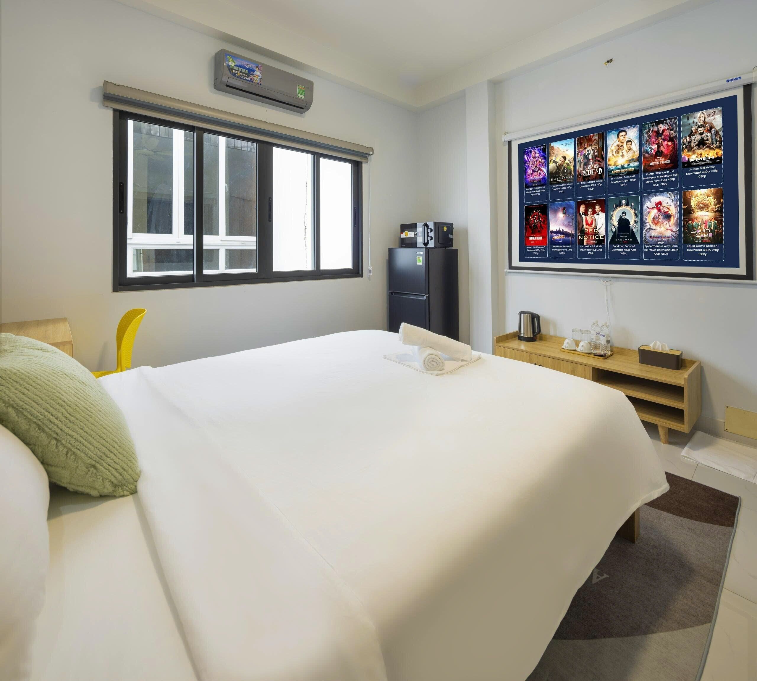A hotel room with a bed, a window, a wall-mounted air conditioner, a refrigerator, a coffee maker, a TV displaying movie posters, and a small wooden table.