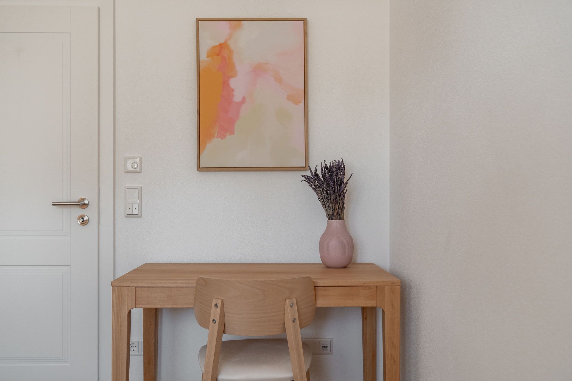 Ein Zimmer mit einem Holztisch, einem Stuhl, einem gerahmten abstrakten Gemälde und einer Vase mit Trockenblumen.