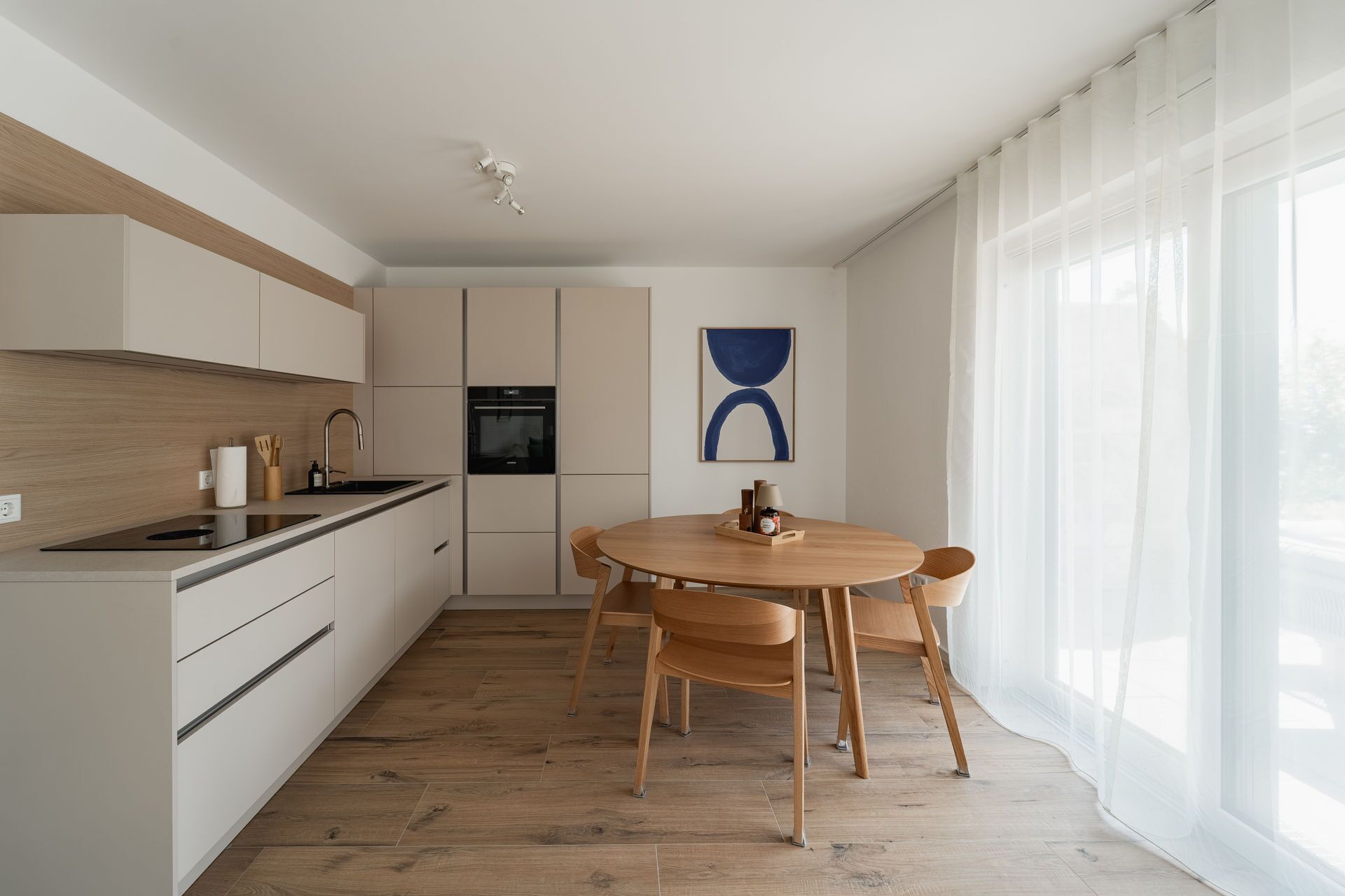 Eine moderne Küche mit Essbereich, Holzboden und einem großen Fenster mit transparenten Vorhängen.