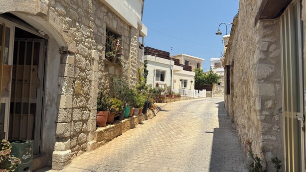 Una estrecha calle adoquinada bordeada por edificios de piedra con balcones y plantas en macetas, bajo un cielo azul despejado.