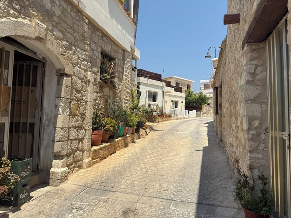 A narrow cobblestone street flanked by stone buildings with balconies, potted plants, and a clear blue sky.