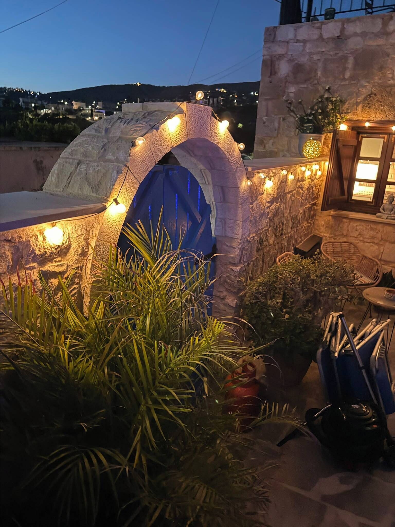 Une arche en pierre surmontée d’une porte bleue, entourée de plantes et illuminée de guirlandes lumineuses.