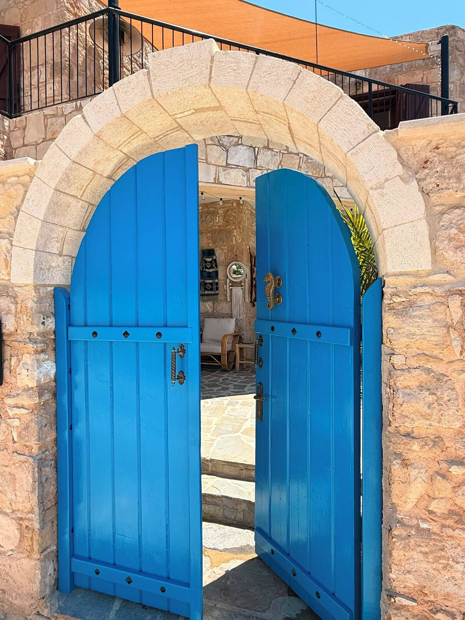 Um arco de pedra com duas portas azuis que dão entrada a um espaço com uma cadeira branca e uma pequena mesa.