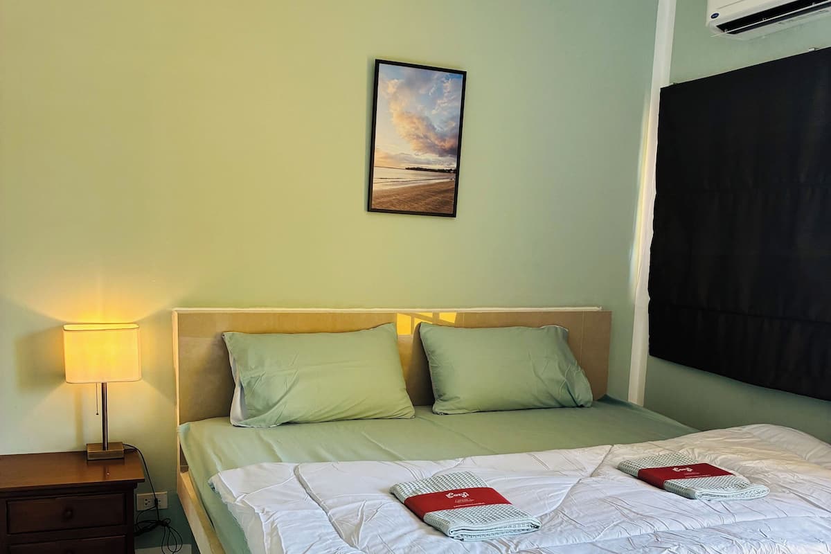 A neatly made bed with a light green bedspread and pillows, a bedside lamp, and a framed picture on the wall.