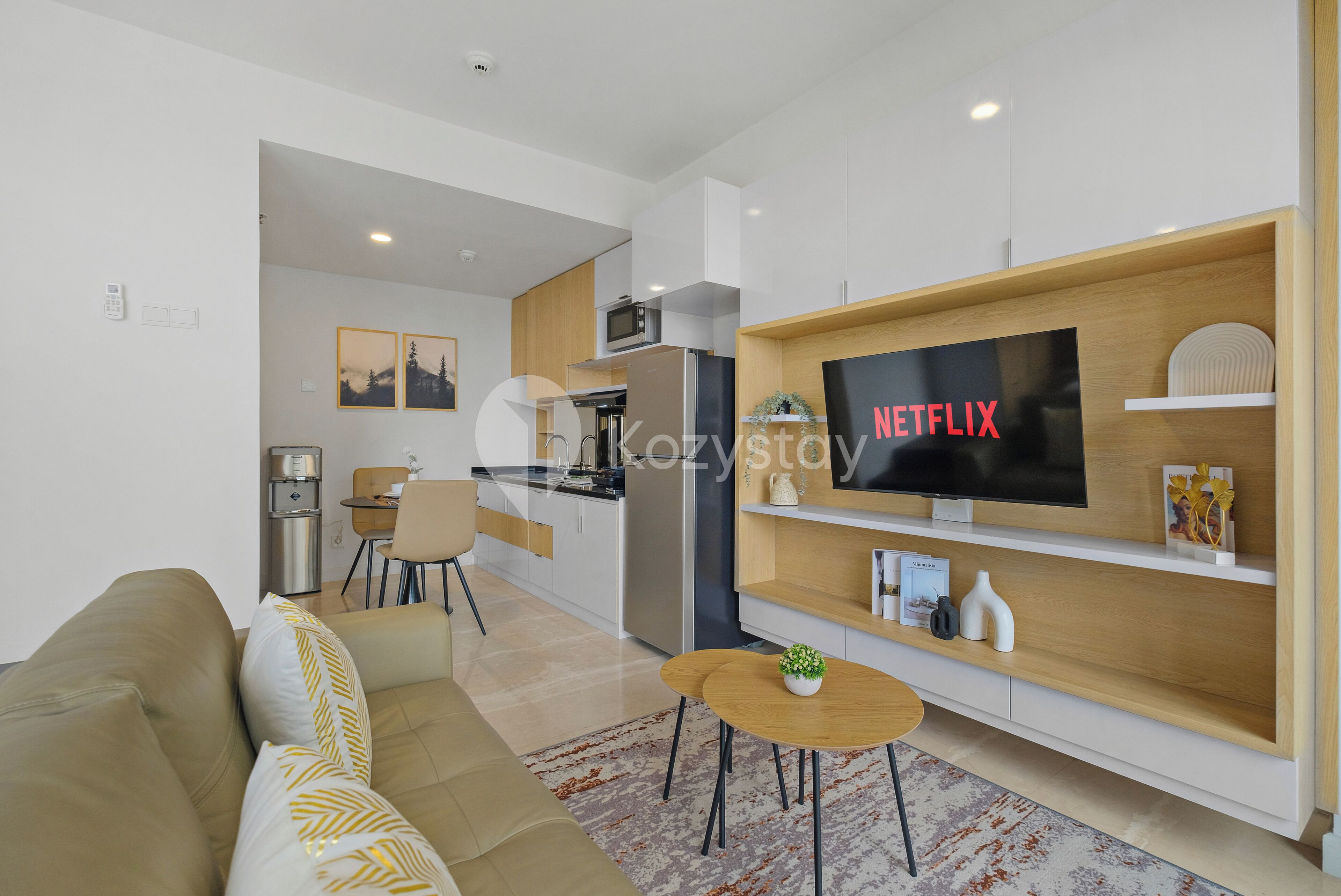 A modern living room with a large flat-screen TV, a sofa, a coffee table, and a kitchen area in the background.