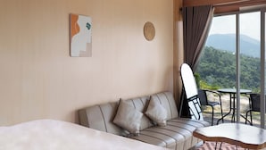 A hotel room with a sofa, a small table, and a view of a mountainous landscape through large windows. - Fortune Glamping (Garung)