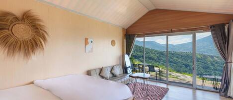 A bedroom with a bed, a sofa, a small table, and a view of the outdoors.